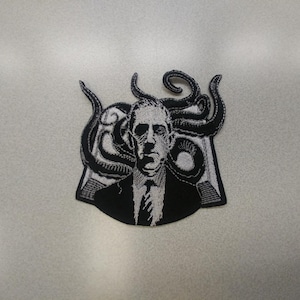 May include: Black and white embroidered patch featuring a man with tentacles around his head. The man is wearing a suit and tie.