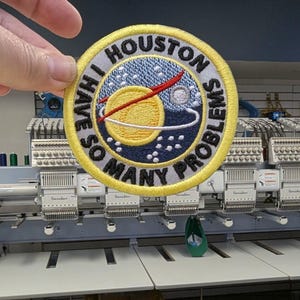 Puede incluir: Parche bordado con el texto "I HAVE SO MANY PROBLEMS" alrededor de un dise&ntilde;o de planeta con anillos y un cometa rojo. El parche tiene un borde amarillo y se sostiene frente a una m&aacute;quina de bordar.