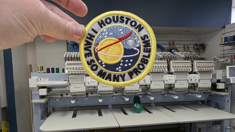 Puede incluir: Parche bordado con el texto "I HAVE SO MANY PROBLEMS" alrededor de un dise&ntilde;o de planeta con anillos y un cometa rojo. El parche tiene un borde amarillo y se sostiene frente a una m&aacute;quina de bordar.