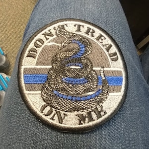 Peut inclure: Un écusson brodé gris et noir avec un serpent à sonnettes enroulé et le texte "Don't Tread On Me". Le serpent est souligné de bleu et il y a une fine ligne bleue au centre de l'écusson.