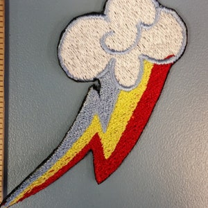 May include: An embroidered iron-on patch featuring a white cloud with a rainbow lightning bolt. The bolt is red, yellow, and blue.