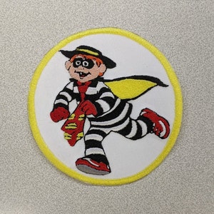 May include: A white embroidered patch with a yellow border, featuring a cartoon character wearing a black and white striped outfit, a red mask, and a yellow cape. The character is running and holding a red and yellow object.