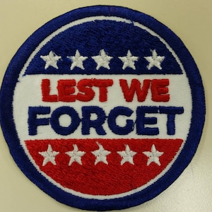 May include: A round, blue, red, and white embroidered patch with the text "LEST WE FORGET" in red and white lettering. There are five white stars above and below the text.