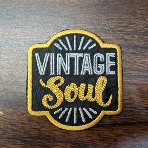 May include: A black embroidered patch with a gold border featuring the text VINTAGE in silver block letters and Soul in gold cursive script. The design includes silver sunburst-style lines above and below the text on a black fabric background.