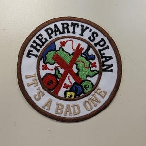 May include: A circular embroidered patch with a brown border and white background. It features a green map with a red X, colorful polyhedral dice, and the black and tan text: THE PARTY'S PLAN IT'S A BAD ONE. The design includes red dotted lines.