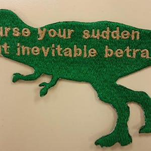 May include: Green embroidered patch shaped like a dinosaur with the text "Curse your sudden but inevitable betrayal" in gold thread.
