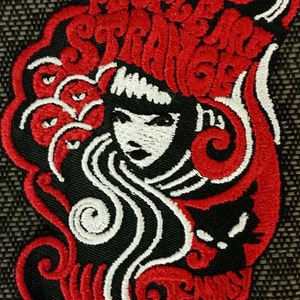 May include: Red and white embroidered patch with a stylized image of a woman with long hair and a cat. The text "Pretty Strange" is embroidered around the woman's head.