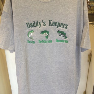 May include: Gray t-shirt with the text "Daddy's Keepers" in green lettering. Below the text are three embroidered fish with the names "Devin", "DeMarion", and "Donovan" underneath each fish.