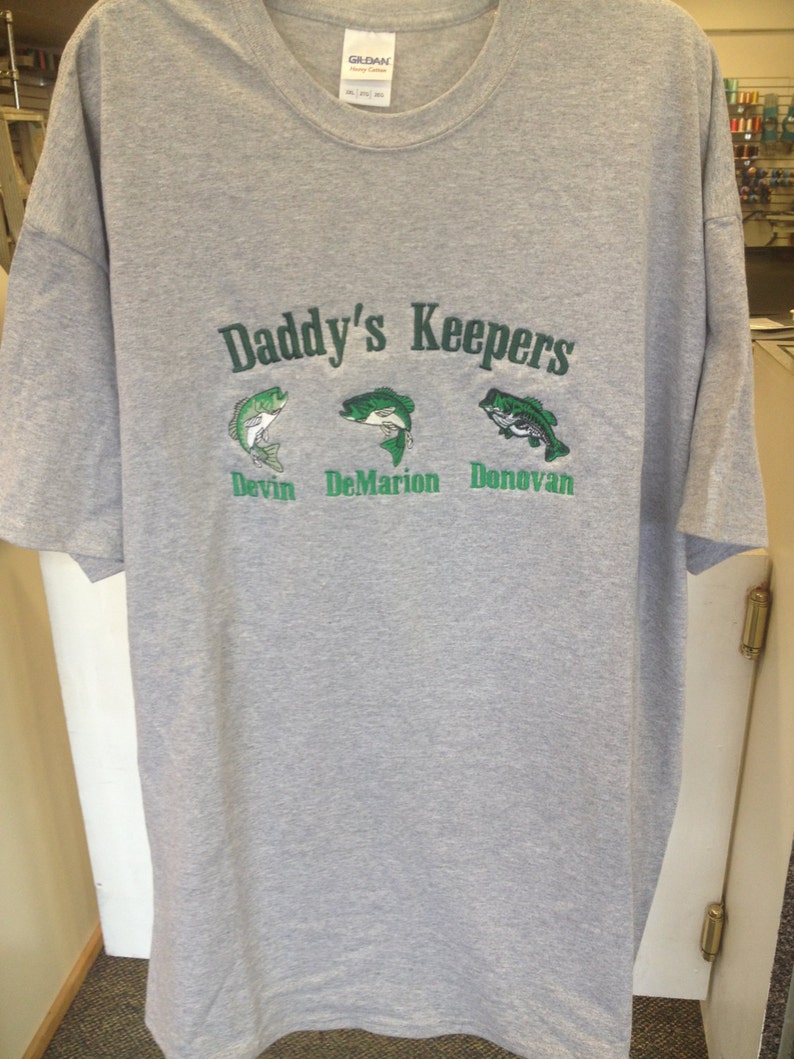 May include: Gray t-shirt with the text "Daddy's Keepers" in green lettering. Below the text are three embroidered fish with the names "Devin", "DeMarion", and "Donovan" underneath each fish.