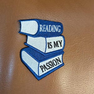 May include: A blue and white embroidered patch featuring three stacked books. The top book reads "READING", the middle book reads "IS MY", and the bottom book reads "PASSION". The patch is on a brown surface.