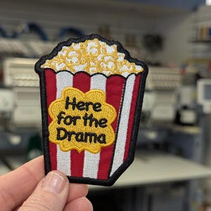 May include: Embroidered popcorn patch with a red and white striped container, topped with yellow popcorn. The patch features the text "Here for the Drama" in a yellow, flower-shaped design. The patch has a black border.