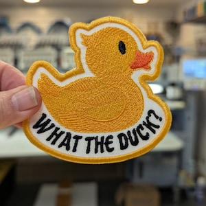 May include: Embroidered patch in the shape of a yellow duck with an orange beak and black eye. The patch has the phrase "WHAT THE DUCK?" in black text on a white oval border.