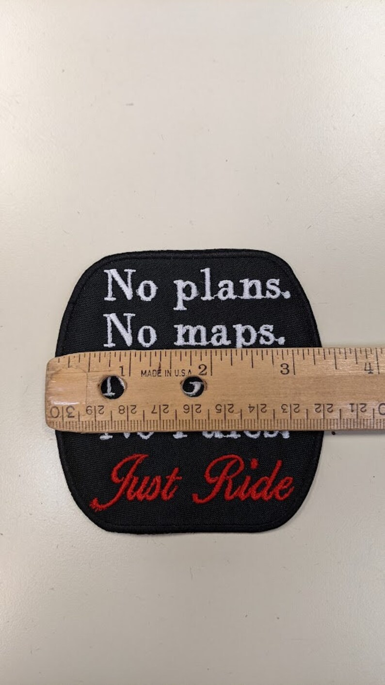 May include: Black embroidered patch with white text that reads "No plans. No maps. Just Ride" in a cursive font. The text "Just Ride" is in red.