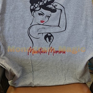 May include: Gray t-shirt with a black and white illustration of a woman flexing her bicep. The woman has red lips and a black ponytail. The text "Mountain Momma" is printed in red below the illustration.