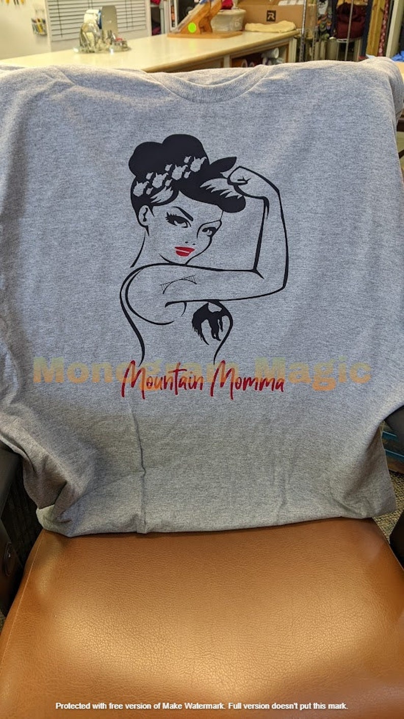 May include: Gray t-shirt with a black and white illustration of a woman flexing her bicep. The woman has red lips and a black ponytail. The text "Mountain Momma" is printed in red below the illustration.