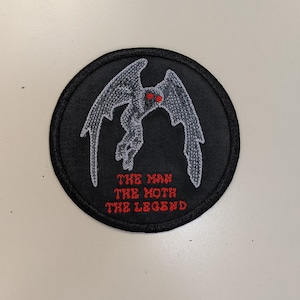 May include: A black and gray embroidered patch with a moth-like creature with red eyes. The text "THE MAN THE MOTH THE LEGEND" is embroidered in red below the creature.