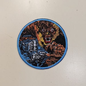 May include: Embroidered patch featuring a snarling, orange and brown werewolf with a blue border. The design includes a detailed illustration of the werewolf's face and clawed hand, with a background of blue water and a white structure.