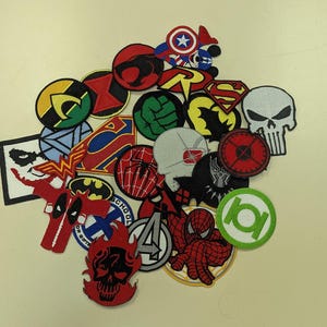 May include: A collection of embroidered patches featuring various superhero and comic book character logos. The patches are in various shapes and colors, including red, blue, green, and yellow. Designs include the Superman logo, Batman symbol, and Spider-Man.