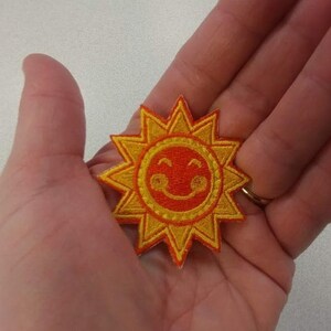 May include: Embroidered patch featuring a smiling sun design. The sun has a red center with a smiling face and yellow rays. This patch is likely for clothing or accessories.