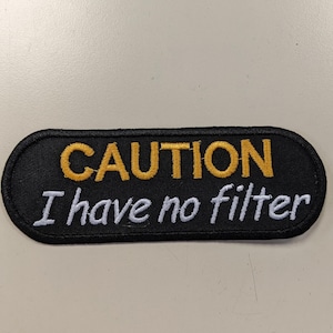 May include: Black oval patch with white and yellow embroidery. The text reads "CAUTION I have no filter".