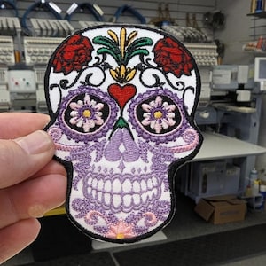 May include: Embroidered skull patch with a black border. The skull features a white background with purple, pink, red, and yellow floral and decorative elements. The patch is held in a hand.