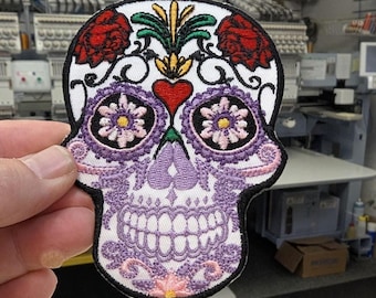 May include: Embroidered skull patch with a black border. The skull features a white background with purple, pink, red, and yellow floral and decorative elements. The patch is held in a hand.