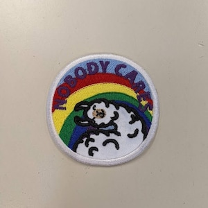 May include: A white embroidered patch with a rainbow and a cartoon sheep. The text "NOBODY CARES" is written in purple around the rainbow.