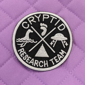 May include: Black and white embroidered patch with the text "Cryptid Research Team" and images of a footprint, a sea monster, a UFO, and a triangle with a circle inside.