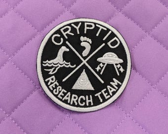 May include: Black and white embroidered patch with the text "Cryptid Research Team" and images of a footprint, a sea monster, a UFO, and a triangle with a circle inside.