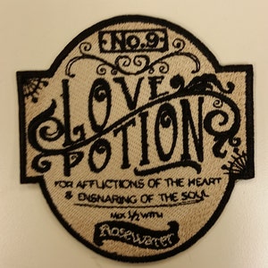 May include: A beige embroidered patch with black stitching. The patch is shaped like a rounded rectangle with a pointed top. The text on the patch reads "No. 91 Love Potion For Afflictions of the Heart & Ensnaring of the Soul  Mix 1/2 with Rosewater".