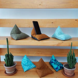 May include: A selection of small, triangular beanbag phone holders in shades of green, brown, and blue. One holder has a black smartphone in it. The beanbags are displayed on wooden shelves with potted cacti.