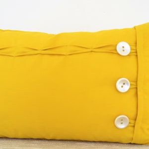 Könnte beinhalten: Ein rechteckiges Kissen in leuchtendem Gelb mit einer Faltenverzierung und drei weißen Knöpfen. Das Kissen ist einfarbig und scheint aus einem weichen Stoff zu sein. Die Knöpfe sind gleichmäßig angeordnet.