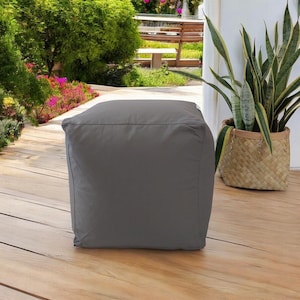 Könnte beinhalten: Ein quadratischer, grauer Outdoor-Hocker. Der Hocker besteht aus einem einfarbigen, glatten Stoff und steht auf einer Holzterrasse. Rechts vom Hocker befindet sich eine Topfpflanze in einem geflochtenen Korb, mit einem Garten im Hintergrund.