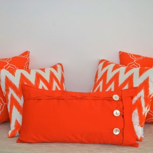 Könnte beinhalten: Eine Sammlung dekorativer Wurfkissen in leuchtendem Orange und Weiß. Die Kissen weisen verschiedene Muster auf, darunter ein geometrisches Gitterdesign und ein Zickzackmuster. Ein rechteckiges Kissen hat Knöpfe.