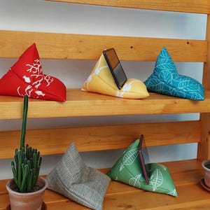 May include: A selection of fabric phone stands in various colours and patterns. The stands are triangular in shape. Designs include a red stand with a white floral pattern, a yellow stand, a teal stand with white leaves, a grey stand, and a green stand with white leaves.