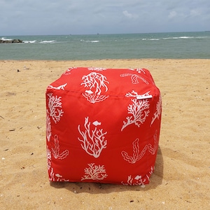 May include: A red square beanbag chair with a white coral and fish pattern sits on a sandy beach. The ocean is visible in the background. The beanbag is a cube shape and appears to be made of a durable fabric.