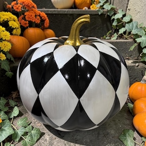 May include: A decorative pumpkin featuring a high-gloss black and white harlequin diamond pattern across its surface. The pumpkin has a smooth, rounded shape and is topped with a metallic gold-painted stem that contrasts with the monochrome body.