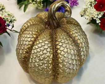 May include: A decorative, gold-colored pumpkin with a textured, scale-like pattern. The pumpkin has a curved stem and is surrounded by colorful flowers. This fall-themed item is ideal for home decor.