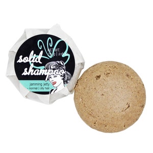 Könnte beinhalten: Ein runder, brauner fester Shampoo-Bar, verpackt in weißes Papier mit einem schwarz-weißen Etikett, auf dem "festes Shampoo" und "Jamming Jetty - normal | fettiges Haar" steht.