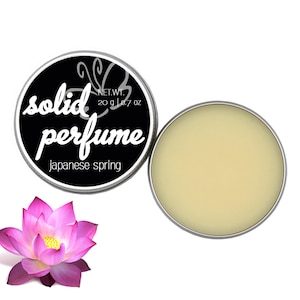 Könnte beinhalten: Eine runde, silberne Dose mit festem Parfüm und einem schwarzen Etikett. Auf dem Etikett steht "solid perfume Japanese spring" in weißer Schrift. Das Parfüm ist hellgelb. Eine rosa und lila Lotusblume ist im Bild.