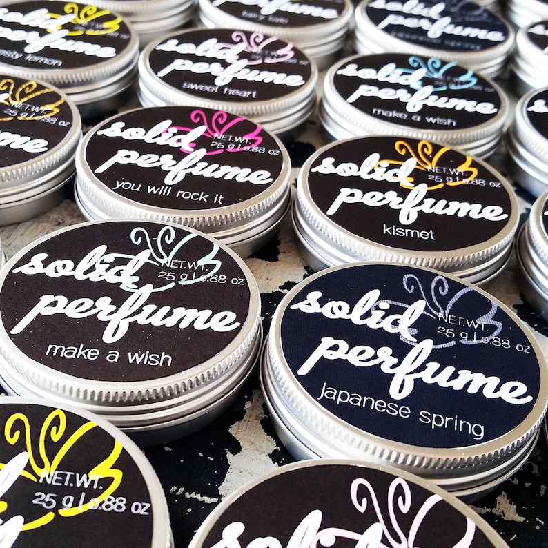 May include: A collection of small, silver tins with black lids. Each tin has a white butterfly design and the words "solid perfume" printed on the lid. The tins contain different scents, including "sweet heart", "you will rock it", "make a wish", "kismet", and "japanese spring".