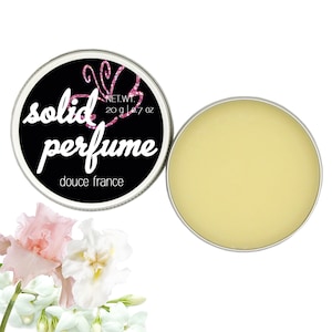 May include: A round tin of solid perfume with a black label that says "solid perfume" and "douce france". The tin is open and the perfume is a pale yellow color. The tin is surrounded by white and pink flowers.