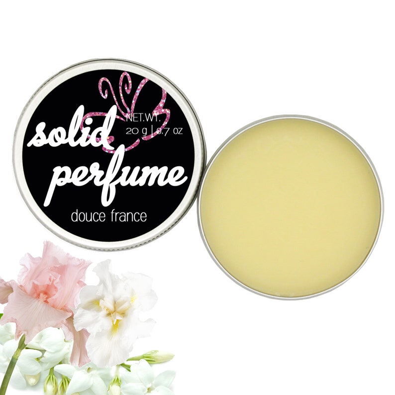 May include: A round tin of solid perfume with a black label that says "solid perfume" and "douce france". The tin is open and the perfume is a pale yellow color. The tin is surrounded by white and pink flowers.