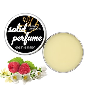 Op de afbeelding: Een ronde blikken doos met een zwart deksel en gouden letters die "solid perfume one in a million" lezen. Het deksel heeft een gouden vlinderontwerp. De doos is open en de binnenkant is gevuld met een lichtgele vaste parfum. De doos staat op een witte achtergrond met een takje witte bloemen en twee rode frambozen.