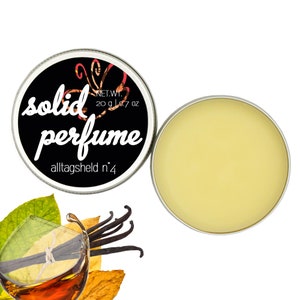Op de afbeelding: Een ronde metalen doos met een zwart deksel en witte letters die "solid perfume alltagsheld n°4" en "NET.WT. 20 g | 0.7 oz" lezen. De doos is open en het deksel is eraf, waardoor een gele vaste parfum zichtbaar is.