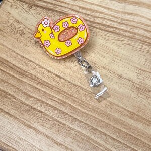 May include: A yellow duck-shaped badge reel with a glittery center and pink floral accents. The badge reel has a clear plastic strap and a metal clip for attaching to clothing or accessories.