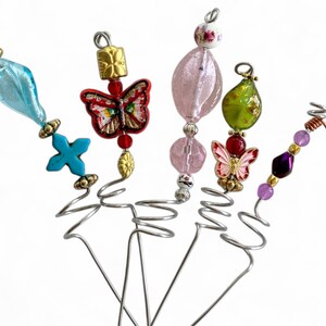 May include: Five decorative wire plant stakes featuring assorted beads and charms. The stakes include elements like a blue glass bead, a red butterfly charm, a pink faceted bead, a green glass bead, and a small iridescent butterfly charm.