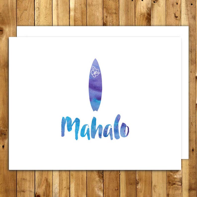 May include: White greeting card with a watercolor design. The card features a blue and purple surfboard with a white flower and the word "Mahalo" in blue watercolor lettering. The card is on a wooden background.