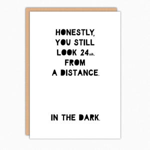 May include: A white greeting card with a brown border. The card features black text that reads: "HONESTLY, YOU STILL LOOK 24ish. FROM A DISTANCE. IN THE DARK."