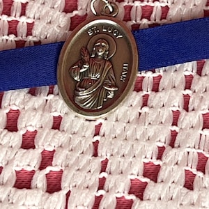 May include: Silver oval-shaped pendant with a depiction of Saint Lucy, a Christian saint, holding a palm branch and a chalice. The pendant has the words "St. Lucy Italy" inscribed on it.